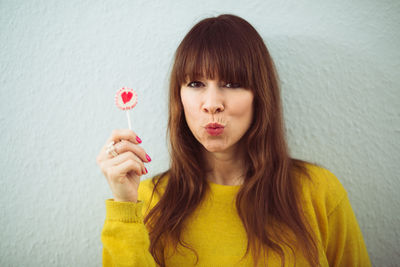 Portrait of redhead woman puckering lips while holding candy against wall