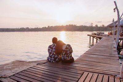 Rear view of couple sitting on pier at lake against sky