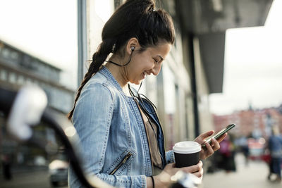 Side view of woman holding smart phone and coffee cup in city