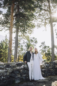 Portrait of newly married couple leaning on retaining wall against forest