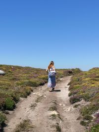 Rear view of woman walking on dirt road against clear blue sky