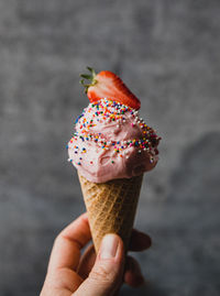 Hand holding strawberry ice cream cone against grey background.