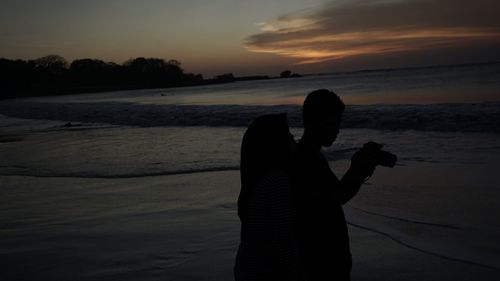 Silhouette man photographing at beach against sky during sunset