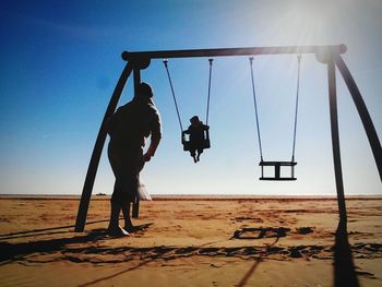 Child swinging by man at beach against sky on sunny day