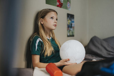 Elementary girl with soccer ball day dreaming while sitting on bed at home