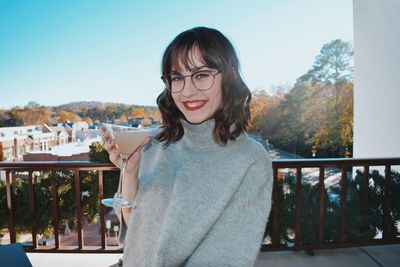 Portrait of smiling young woman holding martini glass while standing against railing