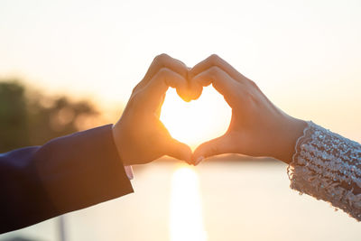 Cropped hand of couple making heart shape against sun during sunset