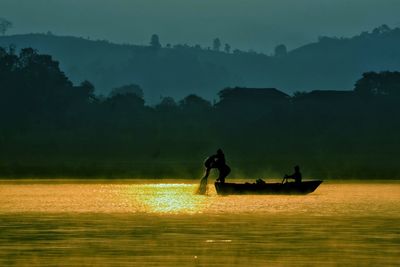 Silhouette men on boat in lake against trees during sunset