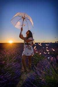 Woman standing by purple flowers on field against sky during sunset