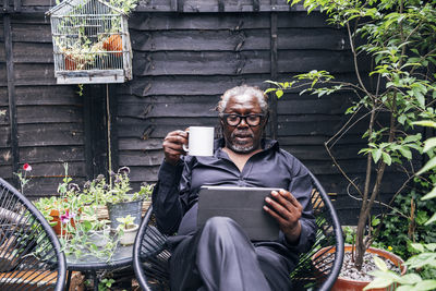 Male professional holding mug looking at digital tablet while sitting on chair in backyard