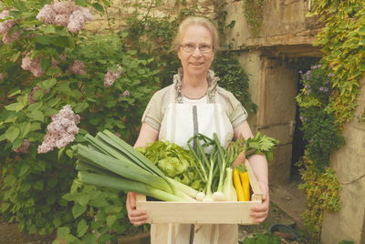 Portrait of smiling senior woman holding fresh vegetables in crate against plants