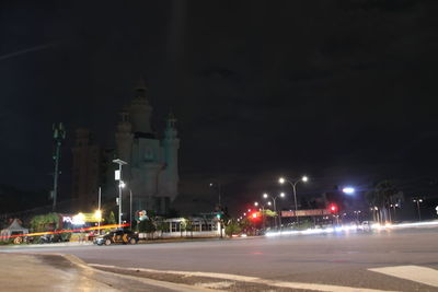 Illuminated street by buildings against sky at night