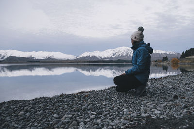 Young woman kneels down at lake tekapo and looks at the southern alps