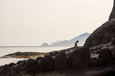 Man standing by rock formation at sea shore against clear sky