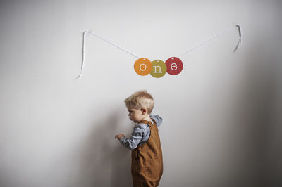 Side view of baby boy standing by text hanging from white wall at home