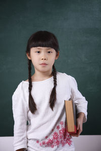 Portrait of girl holding book while standing by blackboard