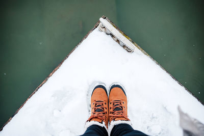 Top view of unrecognizable female feet boots standing on snowy pier by frozen lake during winter.