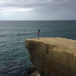 Side view of woman standing on cliff by sea against cloudy sky