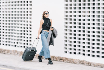 Stylish female tourist with suitcase walking along street in city and looking away