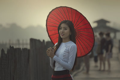 Portrait of young woman with umbrella standing on pier against sky