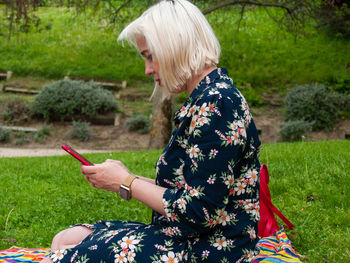 Side view of mature woman using mobile phone while sitting on picnic blanket in park