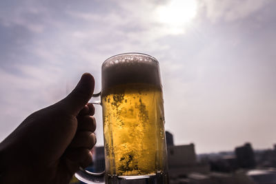 Close-up of hand holding beer glass against sky