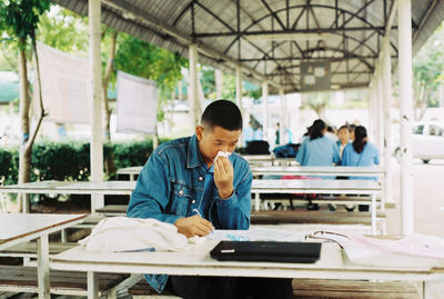 Man cleaning nose with tissue paper while writing over paper on bench