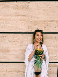 Beautiful woman drinking juice in pineapple while standing against wall