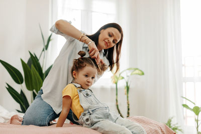 A happy mom combs her little daughter's hair while sitting in bed in the morning