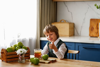 Thoughtful schoolboy is having breakfast with fresh juice and apple waiting for the school bus