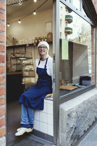 Portrait of smiling senior baker sitting on checkout counter in bakery