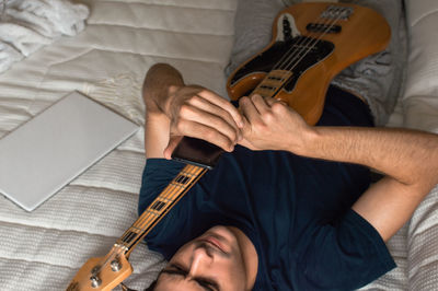 Man lying on a bed using a mobile phone after playing electric bass.