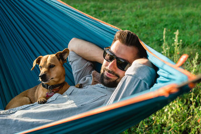 Smiling attractive european man in sunglasses is resting in hammock with his cute little dog.