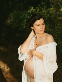 Serene pregnant female in dress touching tummy while standing in countryside dark forest and looking away