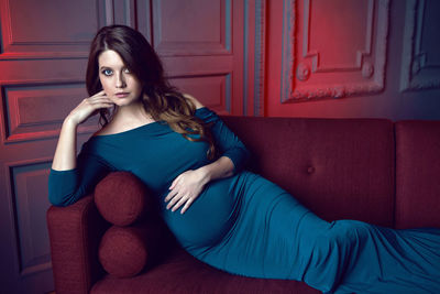 Pregnant woman in blue dress lying on red sofa in studio