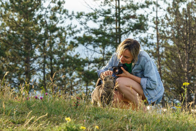 Woman photographer plays with lonely cat walking on forest meadow against blurry green trees