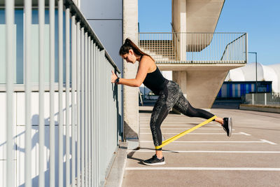 Slim female athlete in sportswear doing exercises with elastic band while standing near metal fence during workout in city resist