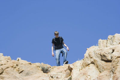 Low angle view of male hiker moving down on rocks against clear blue sky