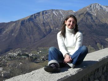 Portrait of smiling woman sitting on retaining wall against mountains