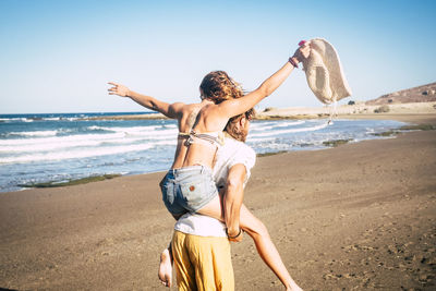 Rear view of man piggybacking woman at beach against clear sky