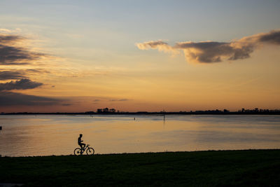 Silhouette person riding bicycle on lake against sky during sunset