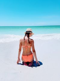 Rear view of teenage girl wearing bikini kneeling at beach against sky during sunny day