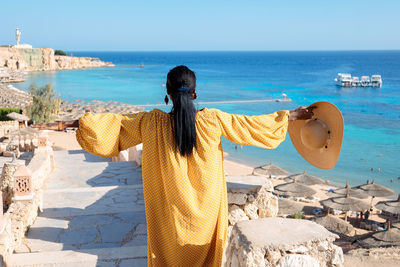 Happy african american woman in yellow dress and sun hat enjoys view of coast of red sea on natural 