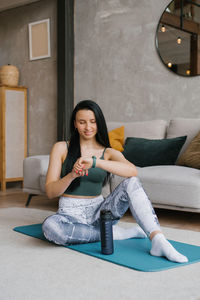 Fitness woman sitting on a mat at home and looking at a wristwatch person