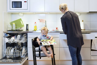 Mother working in kitchen while baby boy sitting on high chair