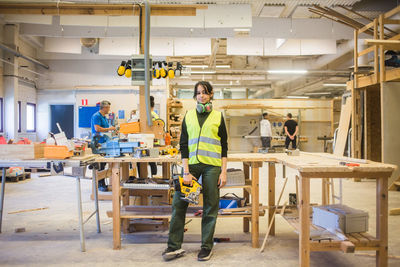 Full length portrait of confident young female trainee holding power tool while standing in workshop