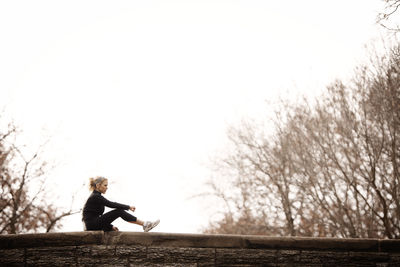 Woman sitting on retaining wall against clear sky