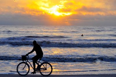 Side view of silhouette woman riding bicycle at beach against cloudy sky during sunset