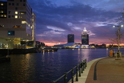 Illuminated buildings by river against sky at dusk