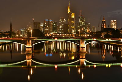 Illuminated bridge over river by buildings against sky at night